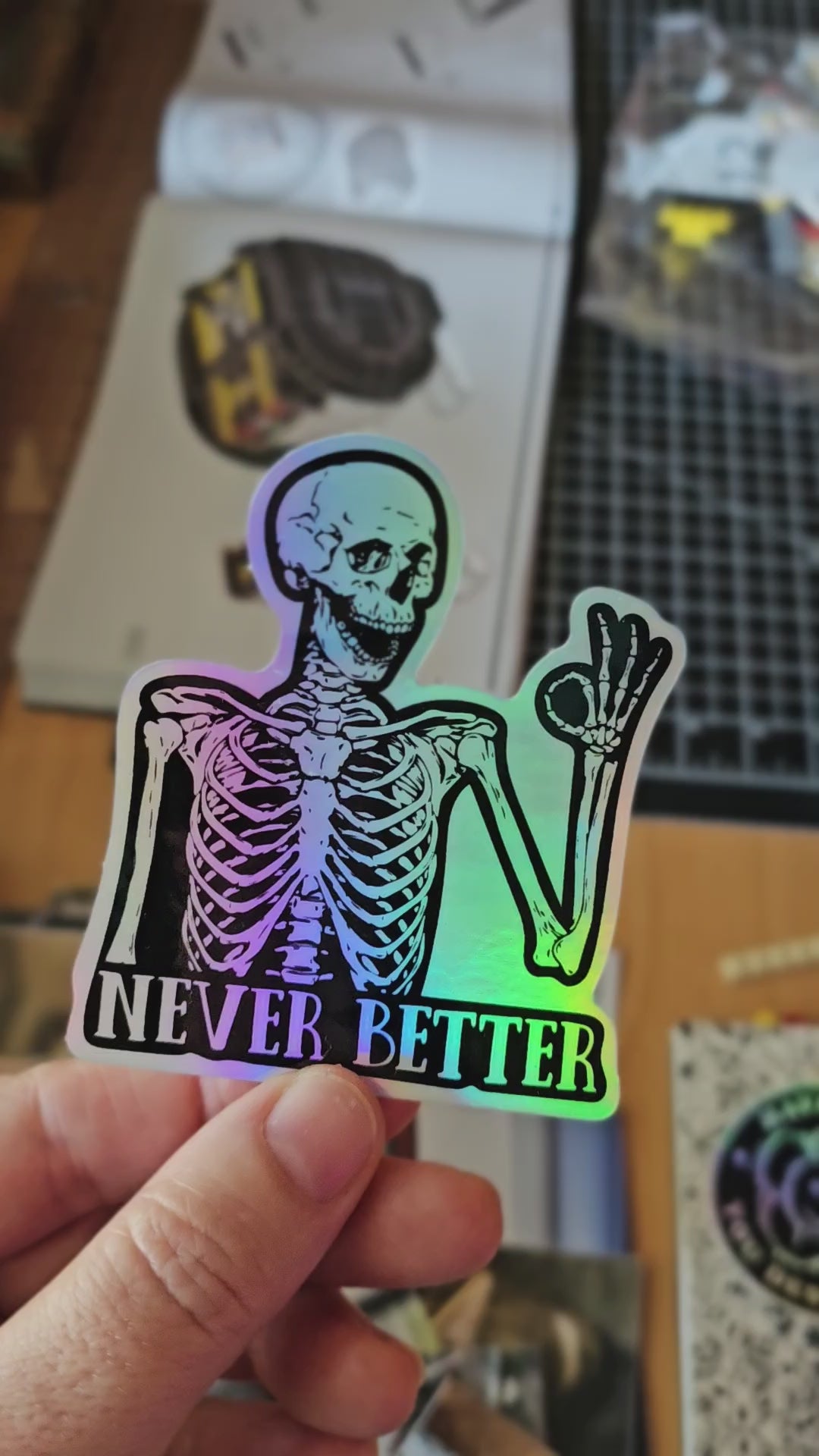 Skeleton sticker with the words “Never Better,” shown in white vinyl and holographic vinyl, featuring a smiling skeleton making an OK hand sign.