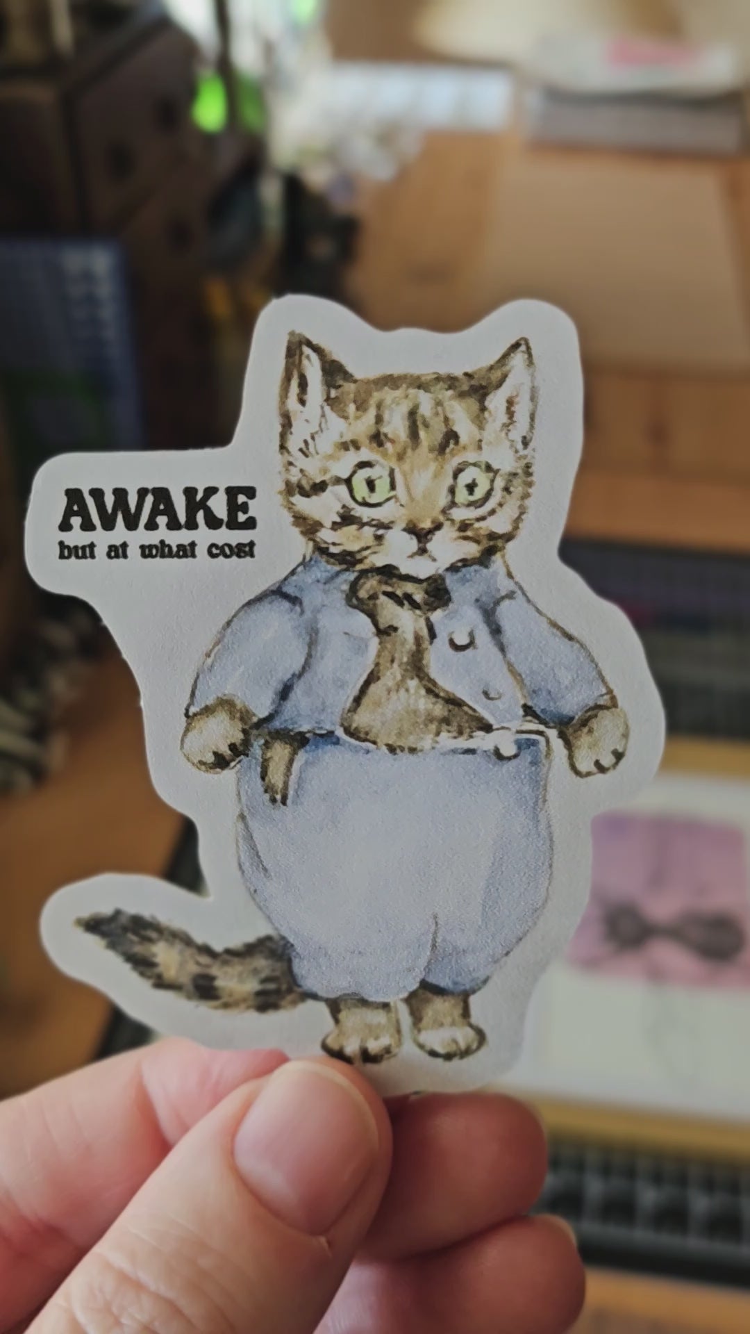 Die cut sticker featuring a vintage storybook-style cat wearing blue clothing with the phrase “Awake but at what cost.”
