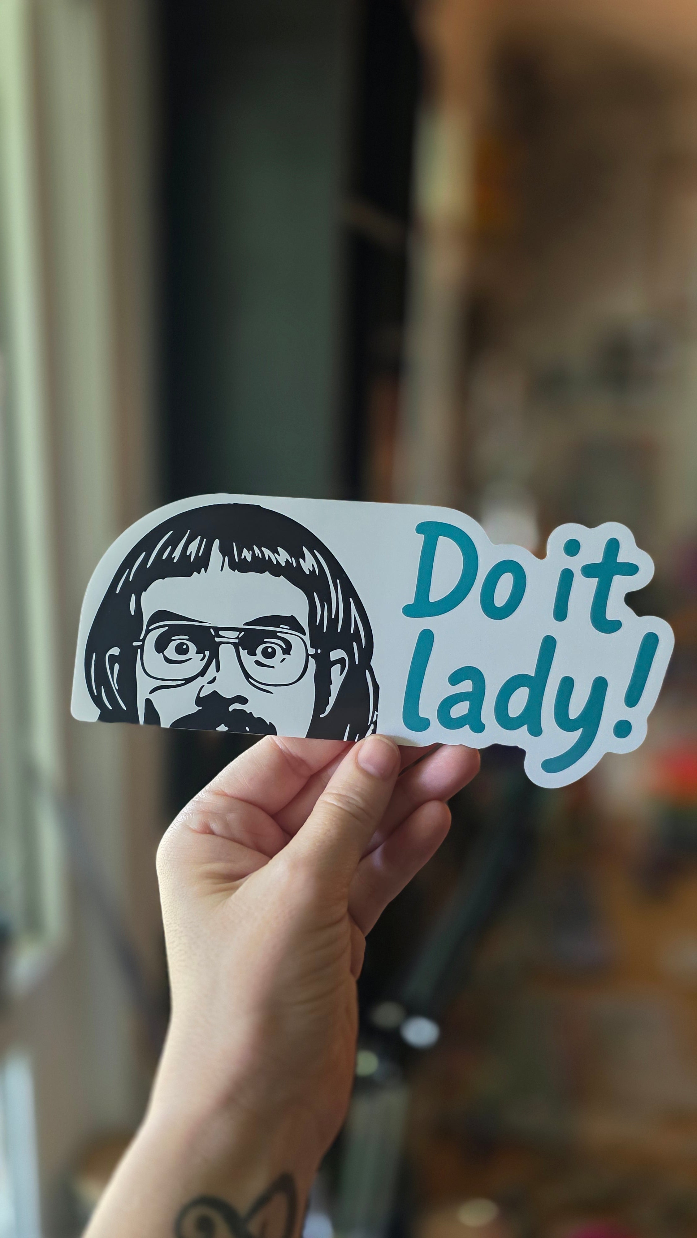 Hand holding a sticker with text and a graphic of a person, against a blurred background.