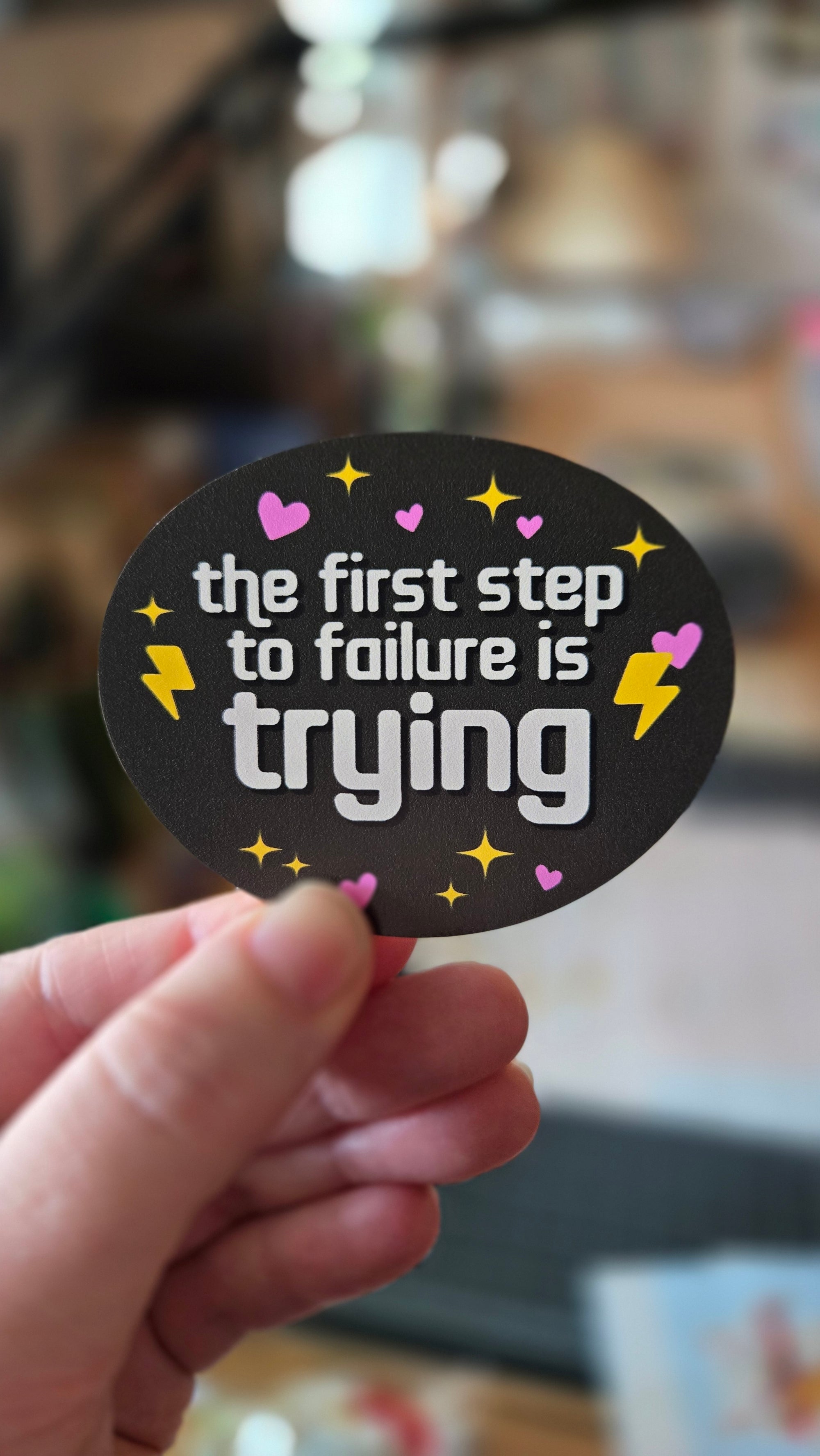 Oval vinyl sticker one black, with the phrase “the first step to failure is trying,” plus hearts, sparkles, and lightning bolts.
