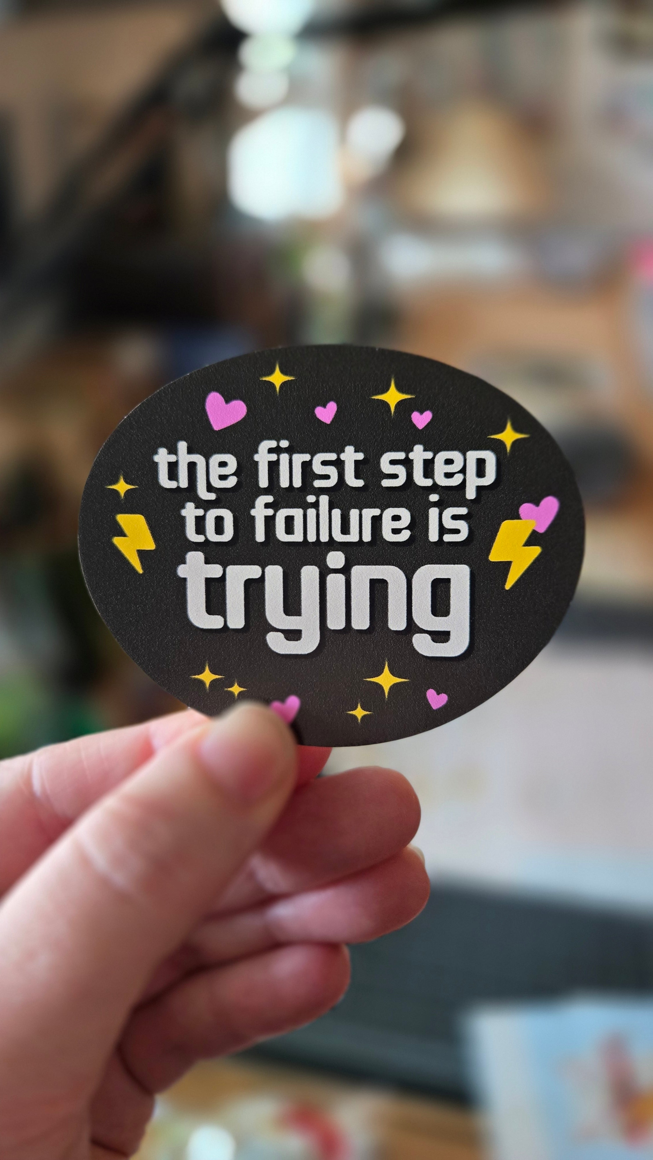 Oval vinyl sticker one black, with the phrase “the first step to failure is trying,” plus hearts, sparkles, and lightning bolts.
