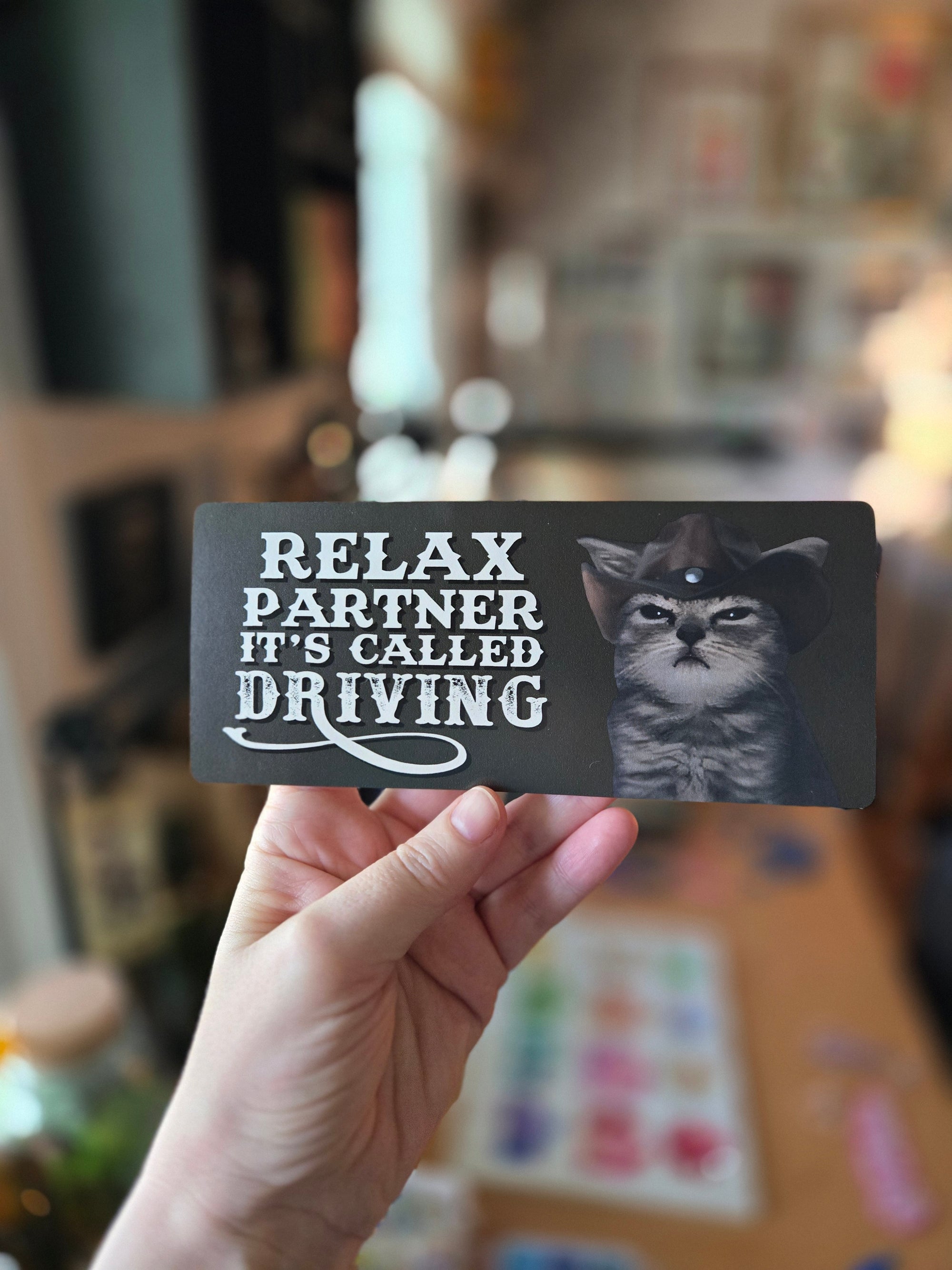 Two rectangular “Relax Partner It’s Called Driving” bumper sticker on dark gray  featuring a serious cowboy cat with bold western-style text.
