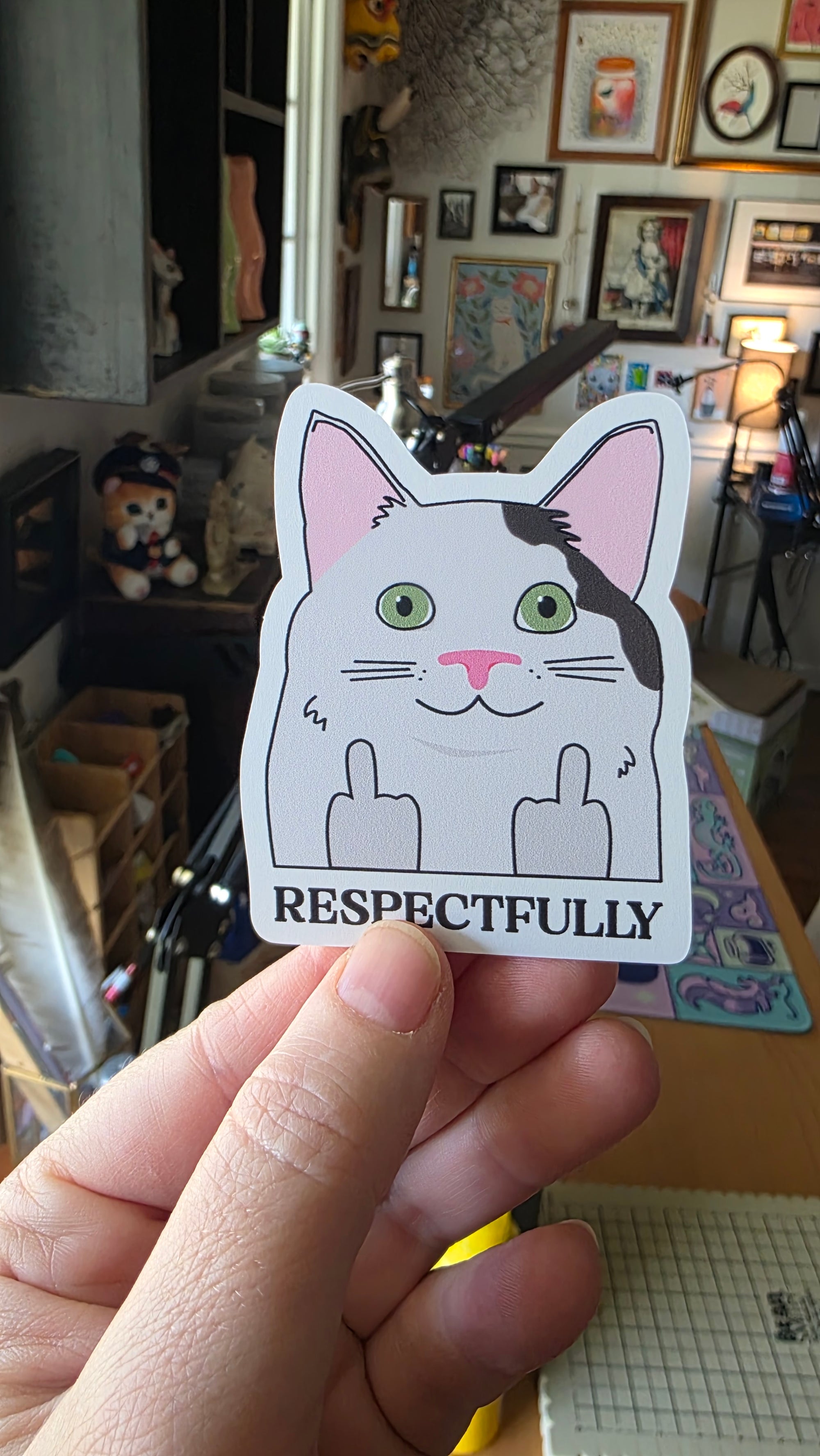 Hand holding a sticker with a cat design and 'Respectfully' text in a casual indoor setting.