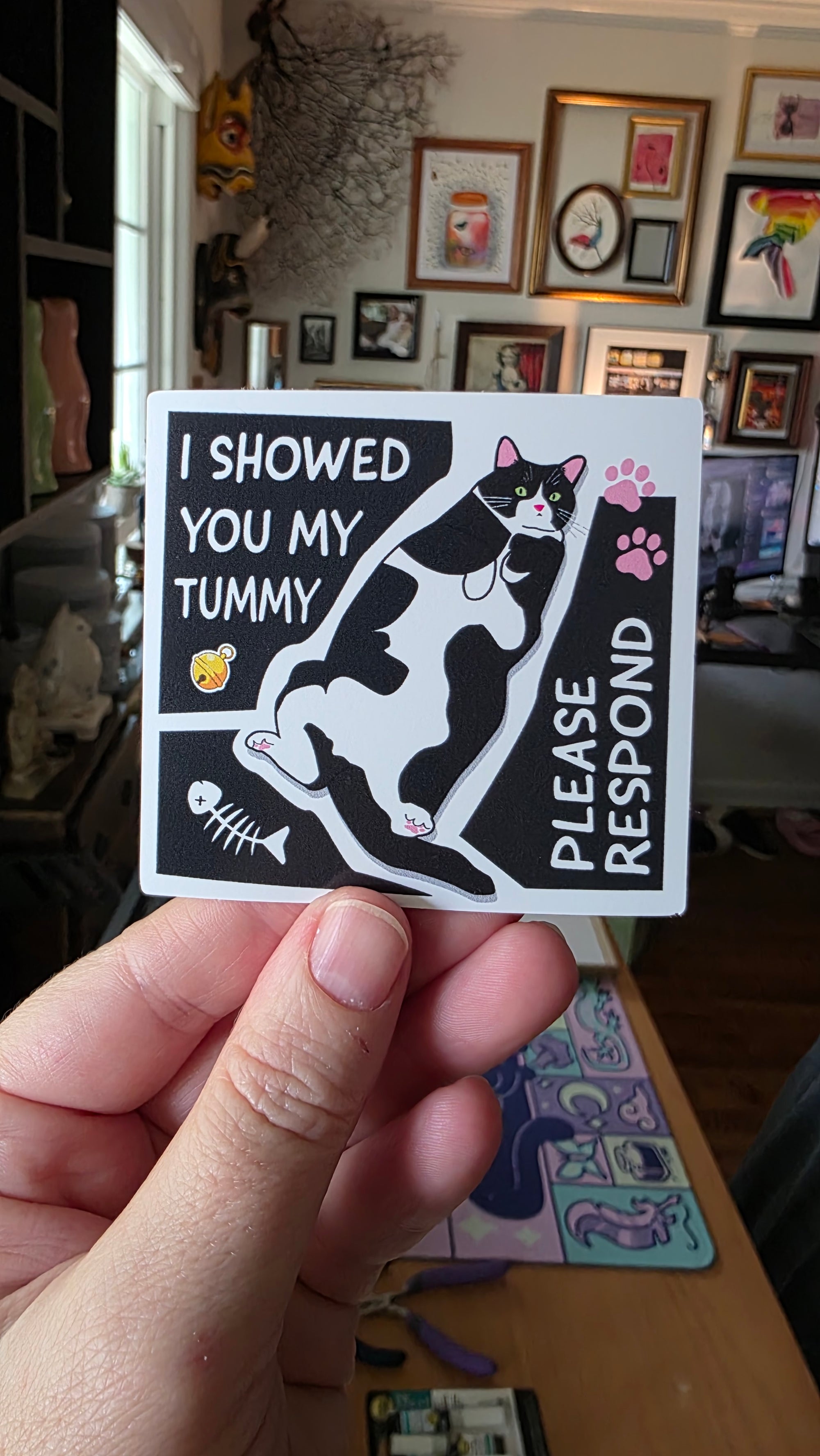 Hand holding a sticker with a cat graphic and text in a casual indoor setting