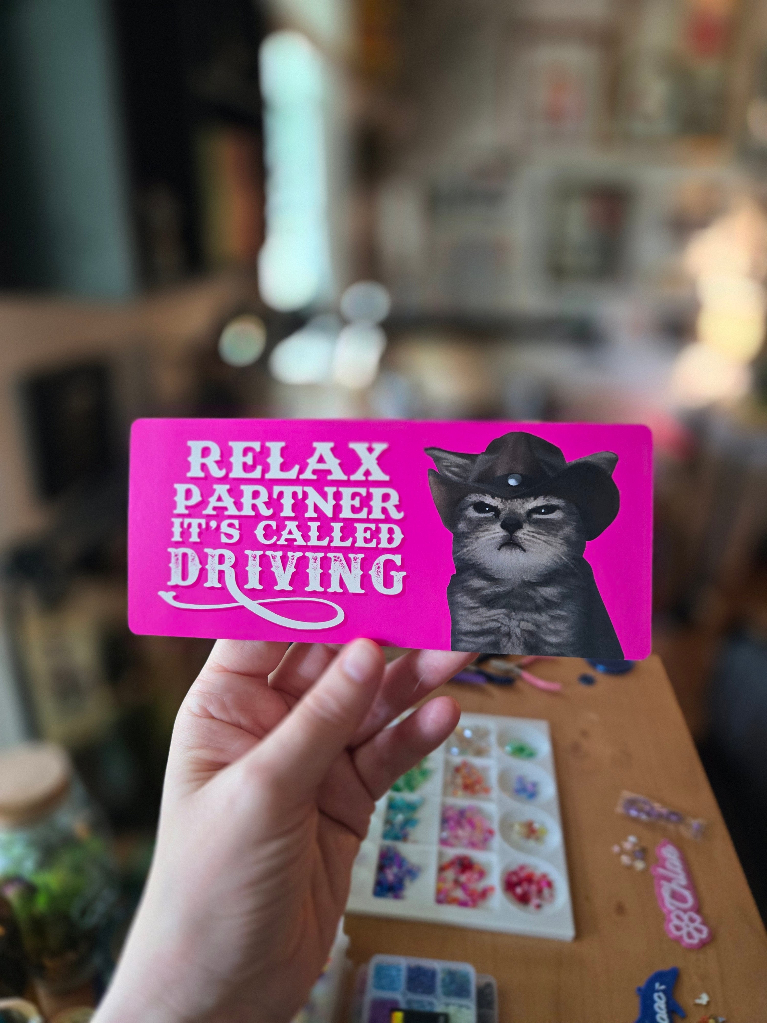 Two rectangular “Relax Partner It’s Called Driving” bumper stickers on pink, featuring a serious cowboy cat with bold western-style text.
