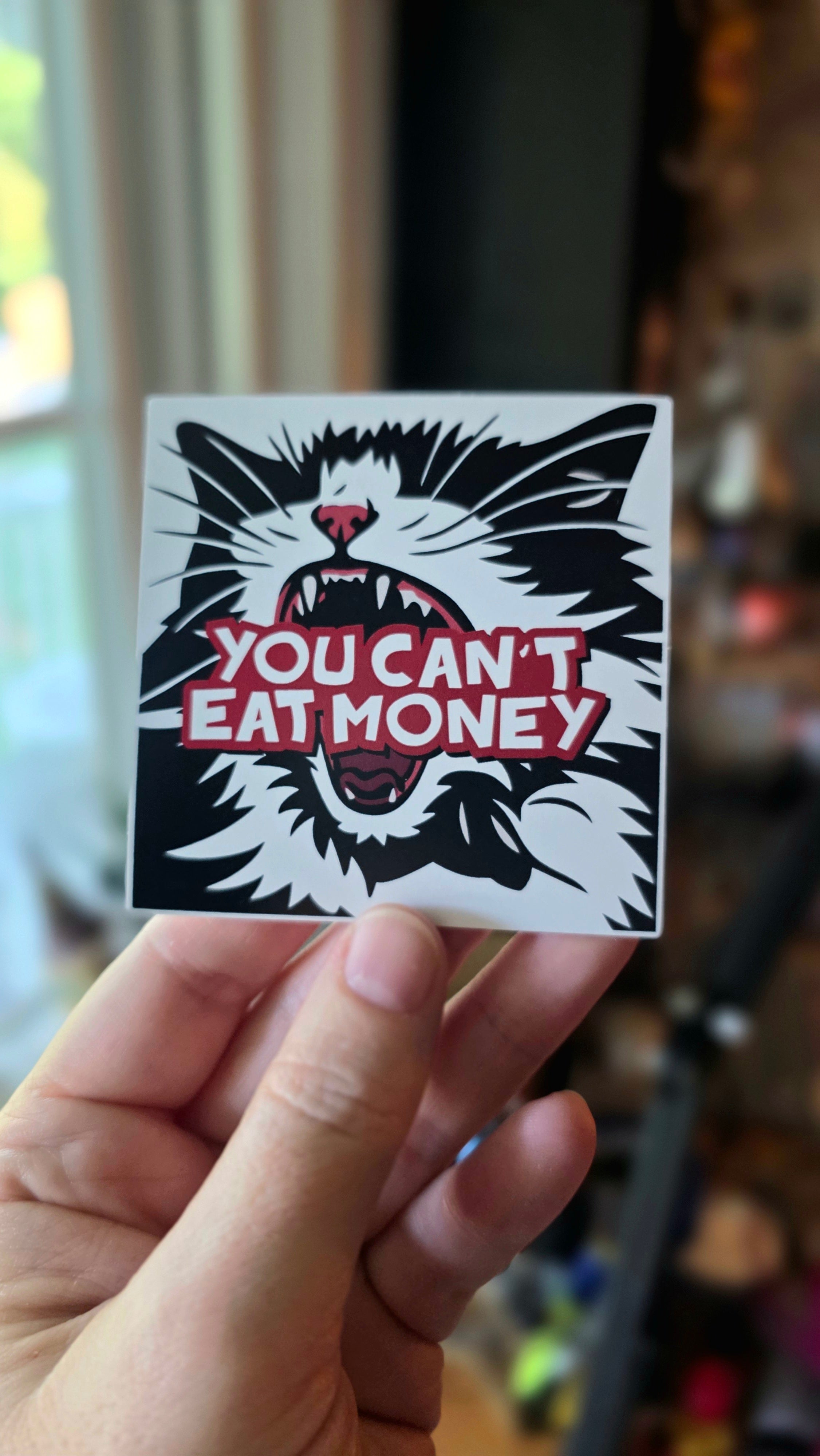 Sticker with a cat graphic and text 'You Can't Eat Money' held by a hand.