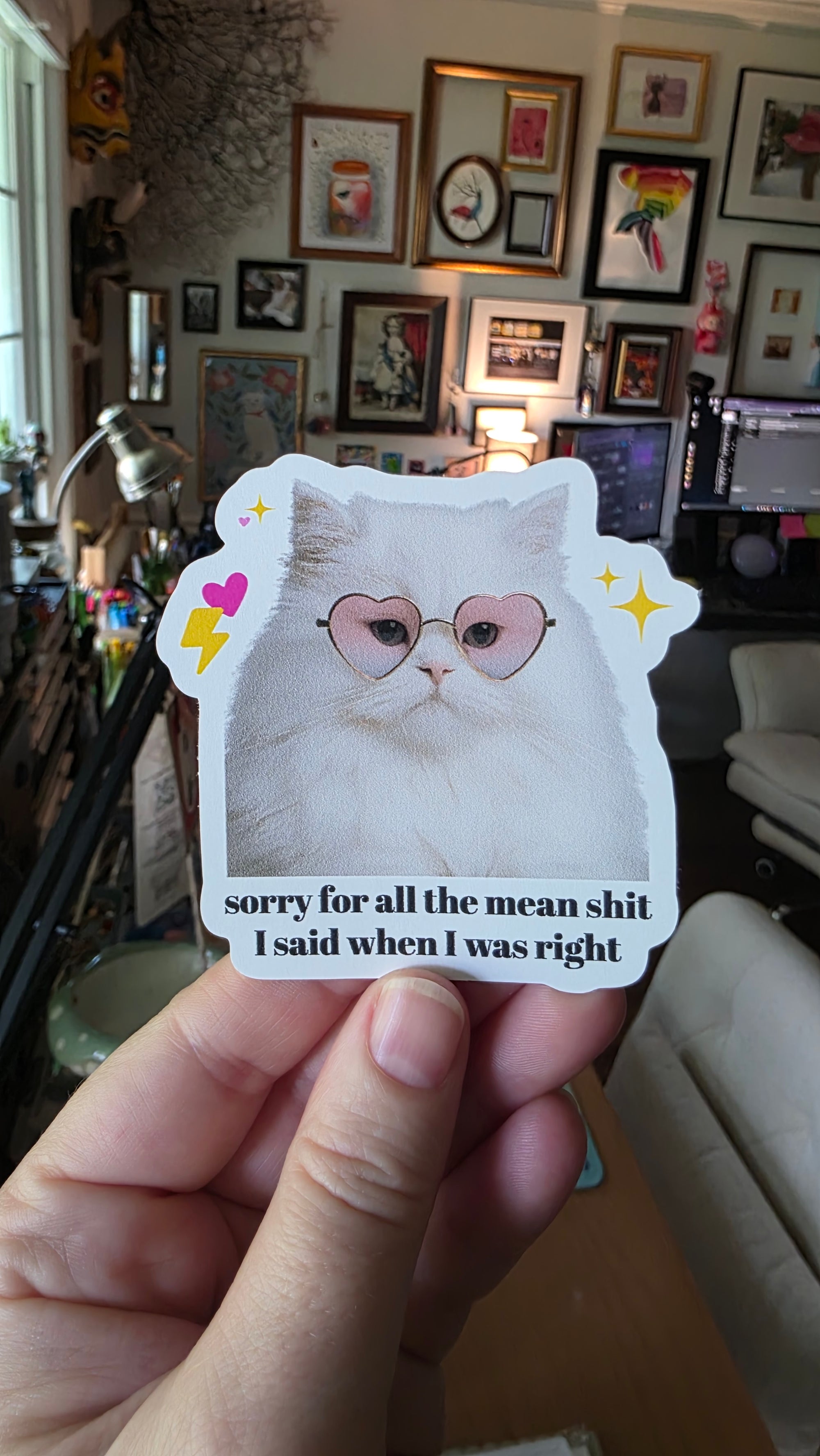 Hand holding a sticker of a cat illustration and text in a room with framed pictures on the wall.
