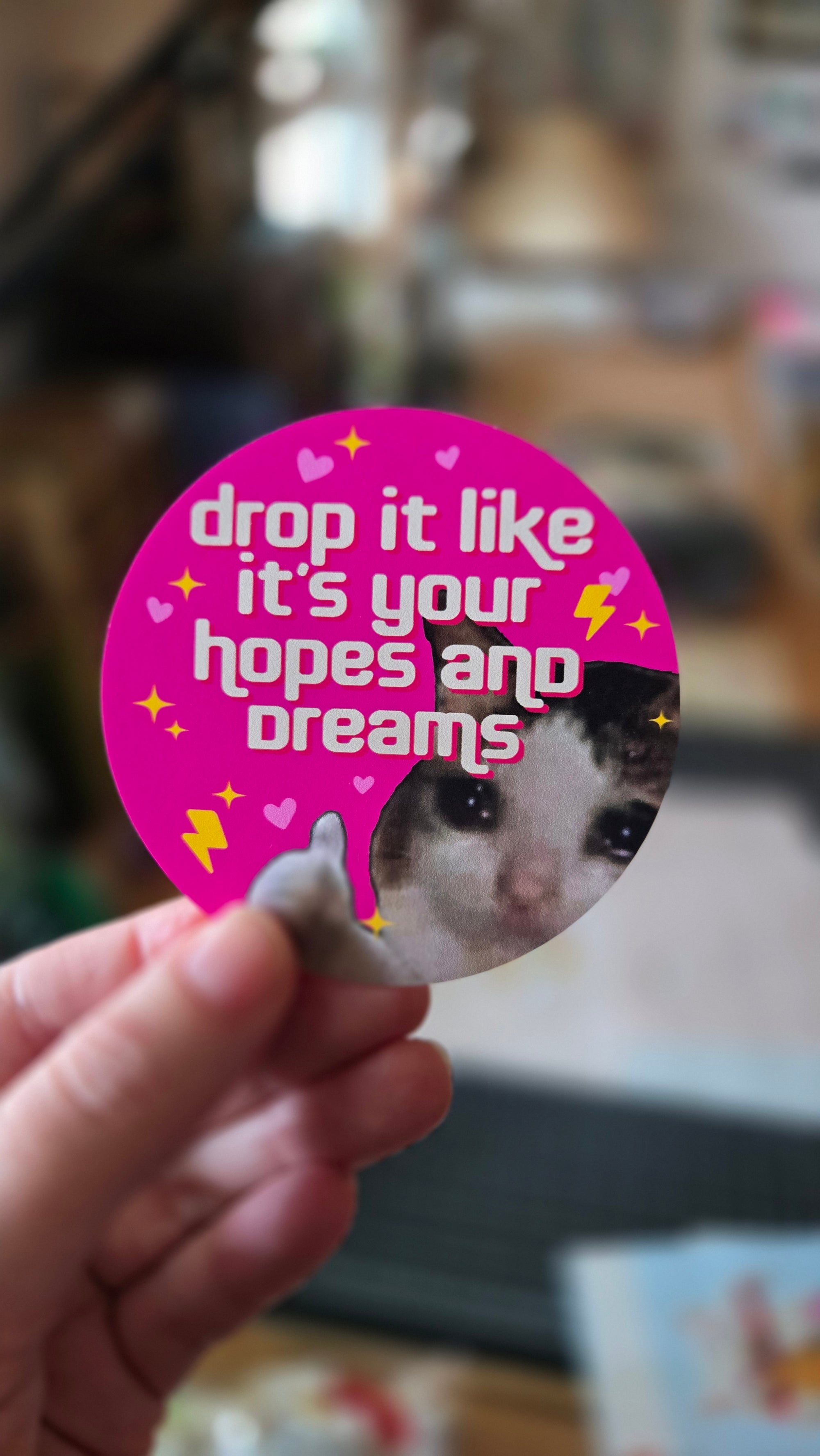 A round sticker with a demotivational phrase 'drop it like it's your hopes and dreams' featuring a sad cat giving a thumbs-up surrounded by sparkles, lightning bolts, and hearts.