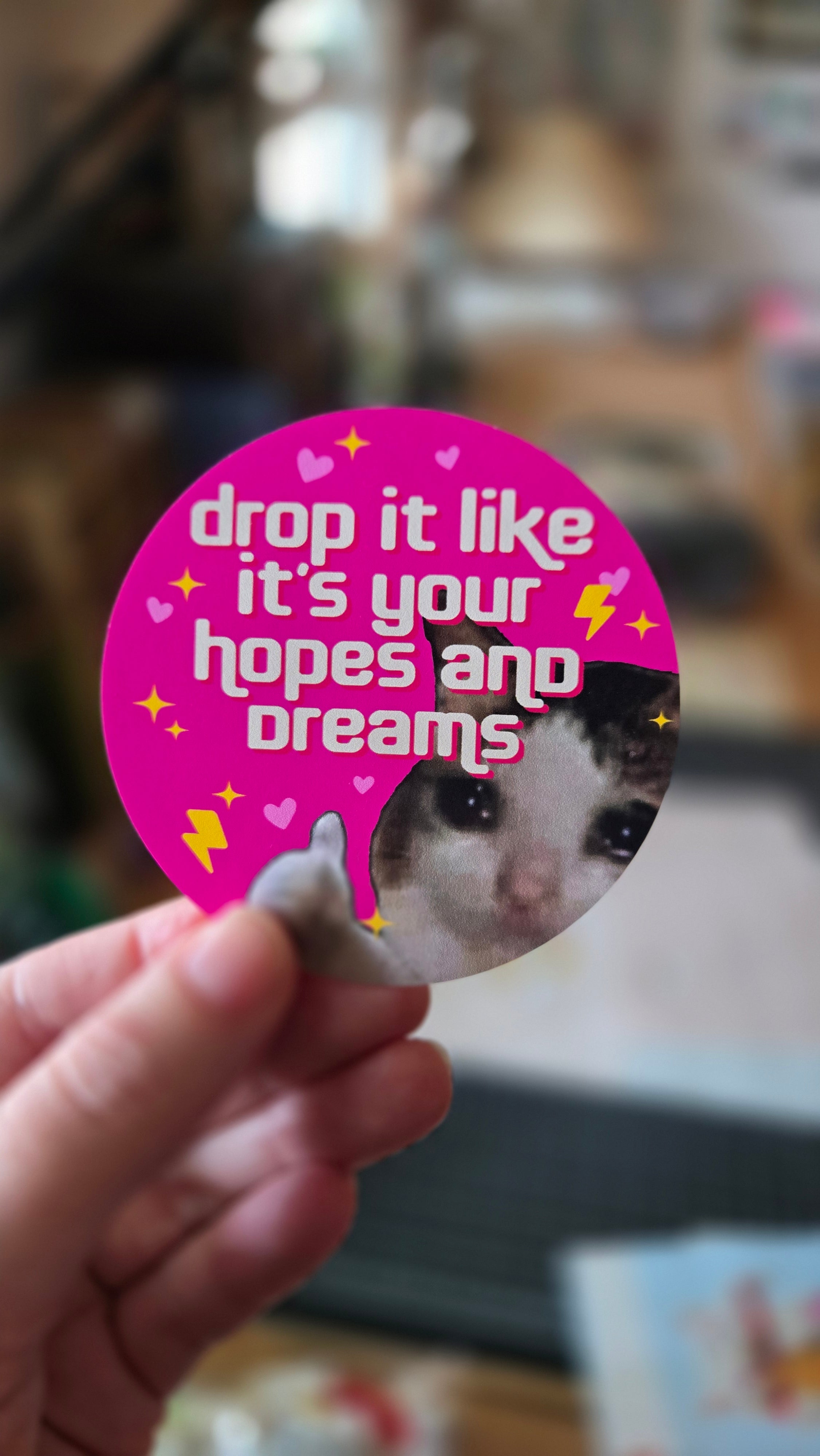 A round sticker with a demotivational phrase 'drop it like it's your hopes and dreams' featuring a sad cat giving a thumbs-up surrounded by sparkles, lightning bolts, and hearts.