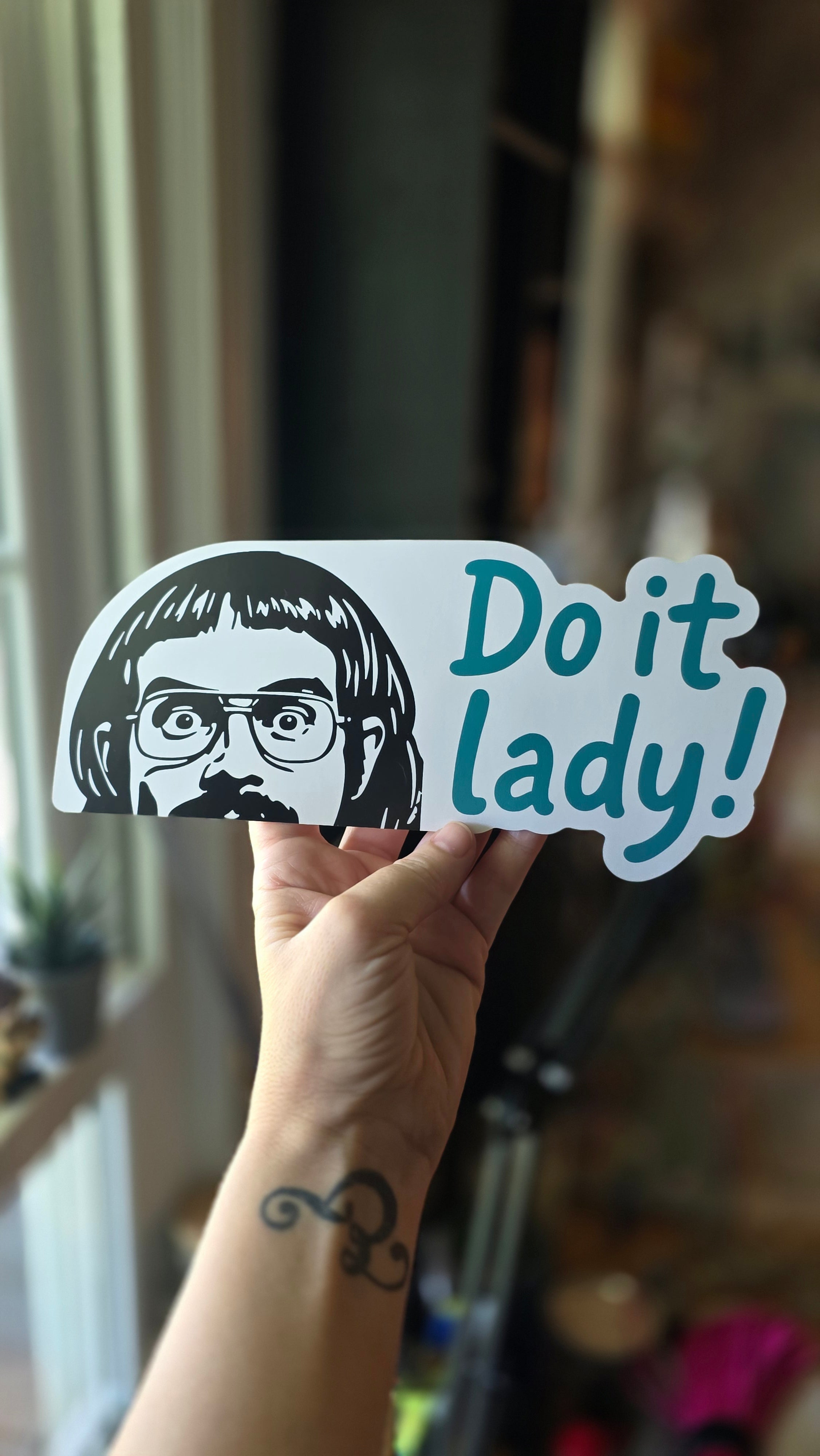 Sticker with 'Do it lady!' text held by a hand with a tattoo.