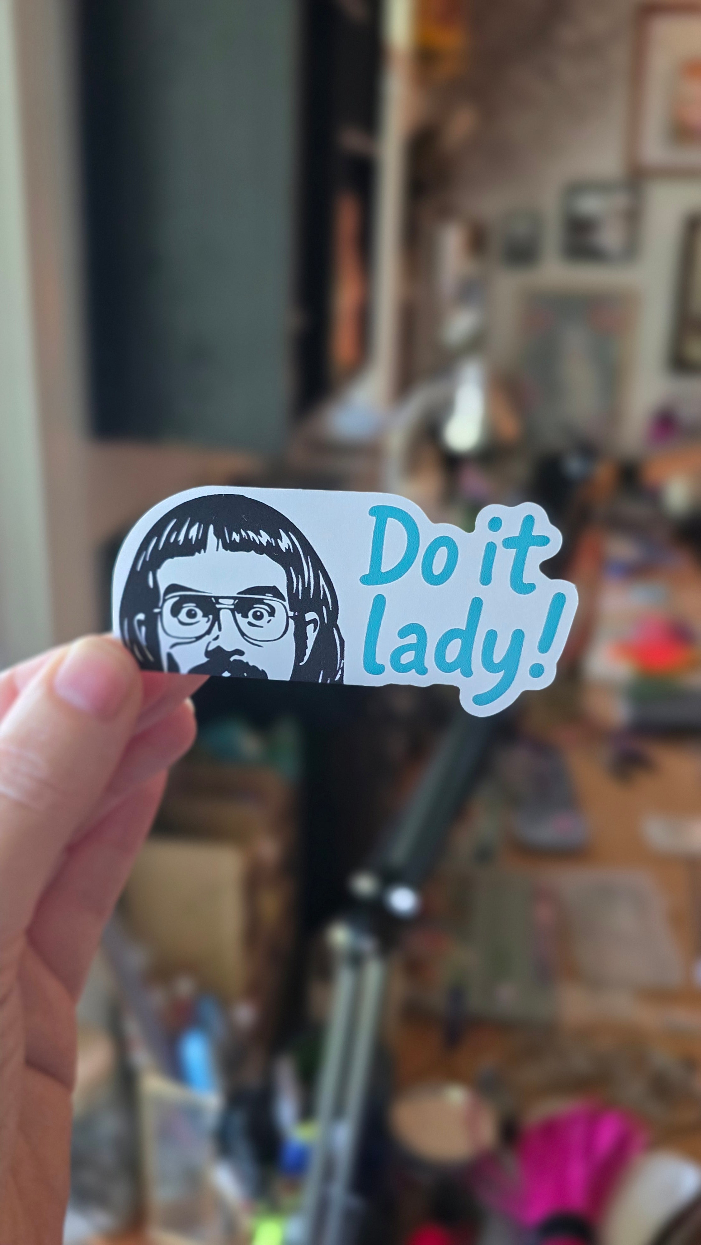 Sticker with 'Do it lady!' text held in front of a blurred indoor background