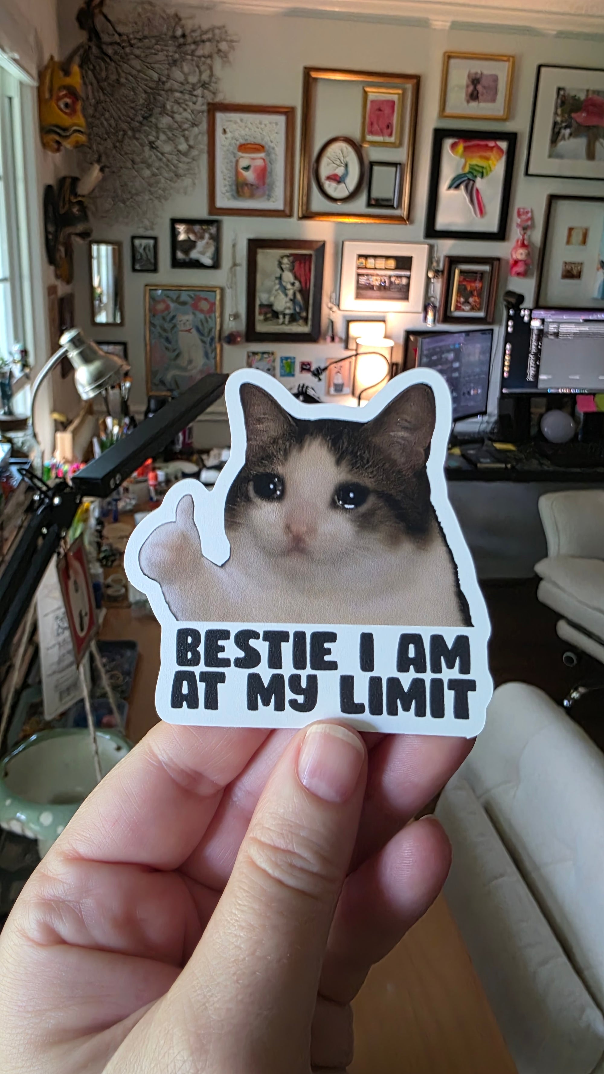 Person holding a sticker with a cat and text 'Bestie I am at my limit' in a room with framed pictures on the wall.