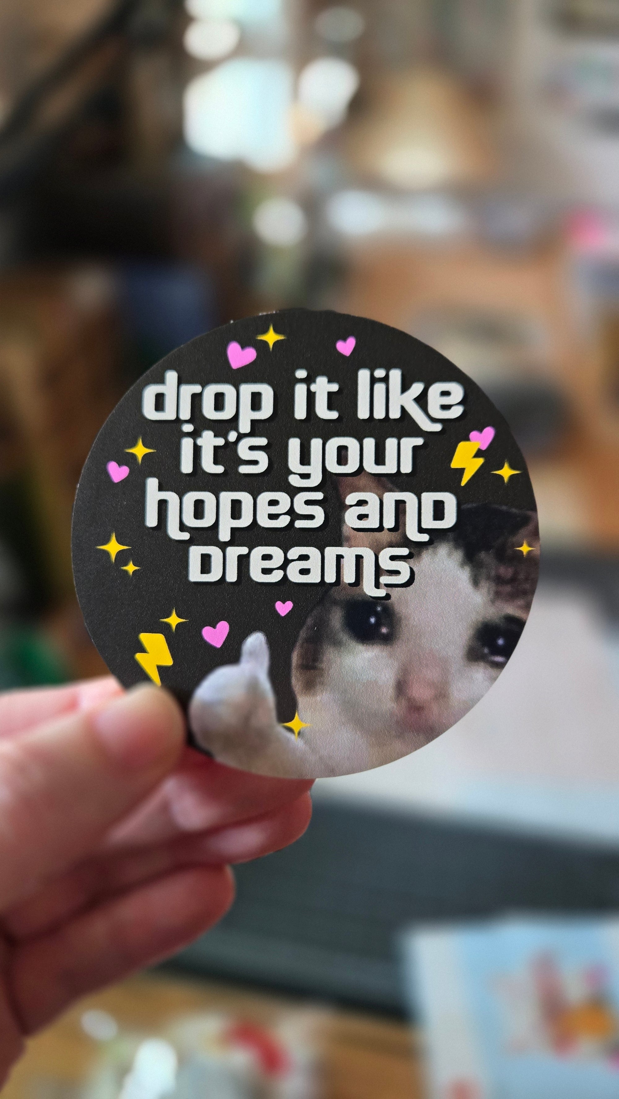 A round sticker with a demotivational phrase 'drop it like it's your hopes and dreams' featuring a sad cat giving a thumbs-up surrounded by sparkles, lightning bolts, and hearts.
