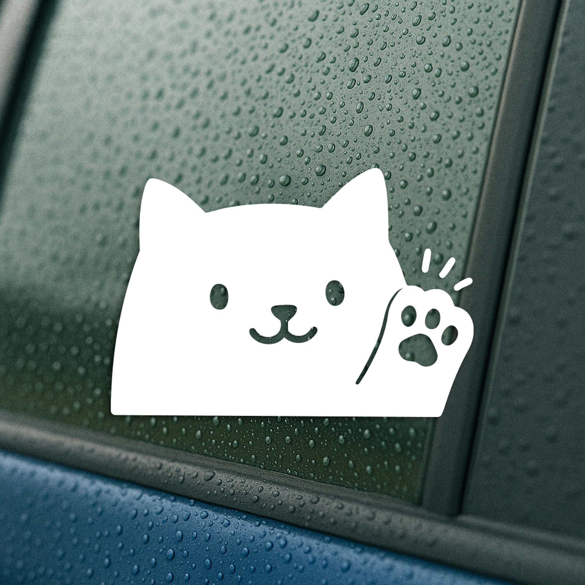 White vinyl decal on a rainy car window featuring a cute peeking cat face with one raised paw.
